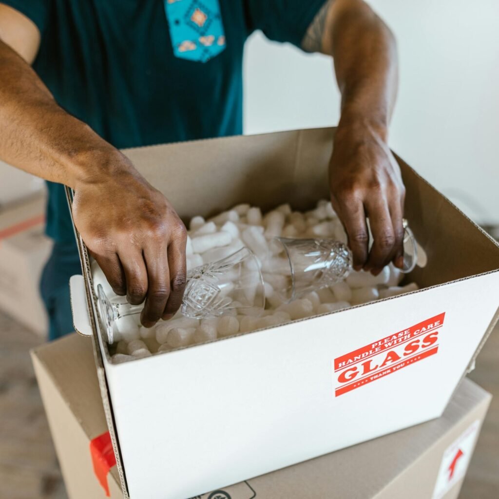 Hands carefully packing glassware into a cardboard box with padding for safe transport.